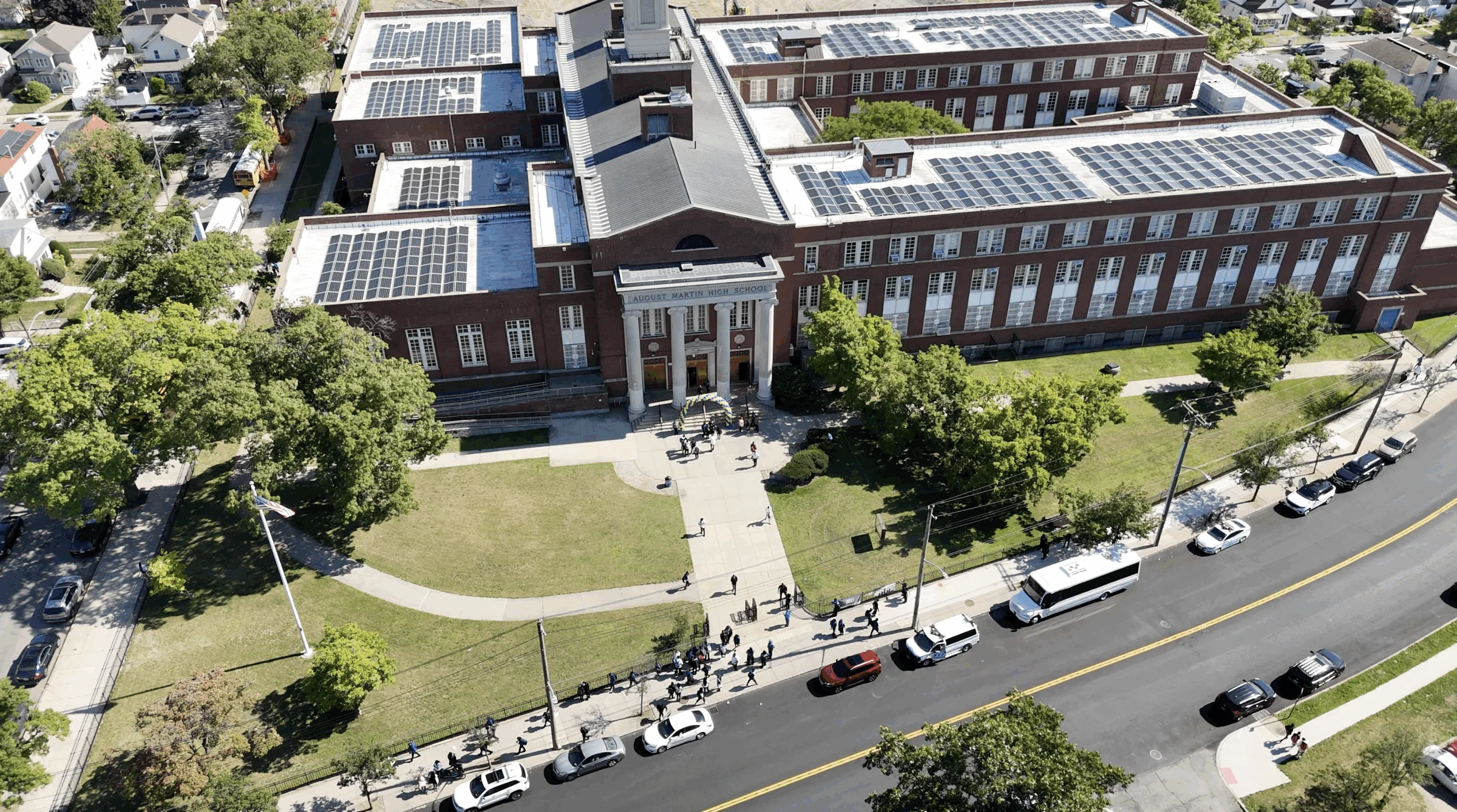 Aerial view of a large brick building with solar panels on the roof, people gathered at the entrance, and cars parked along the street out front.